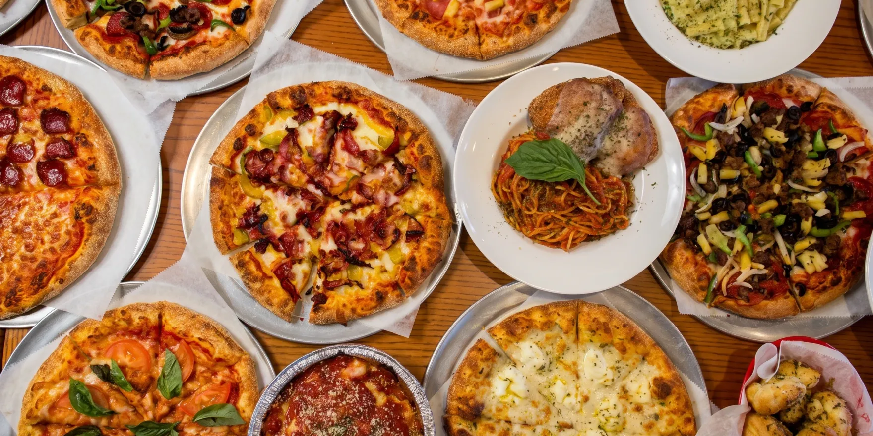 An overhead table covered with Uncle Reds pizzas, pastas, a baked ziti and a basket of garlic knots